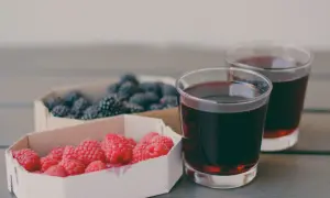 Dwa kieliszki ciemnego soku z jeżyn, obok koszyczki z malinami i jeżynami. Sok z jeżyn właściwości zdrowotne.