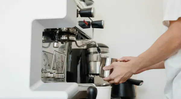 Barista spienia mleko w dzbanku, używając dyszy ekspresu. W tle widoczny blok zaparzający.