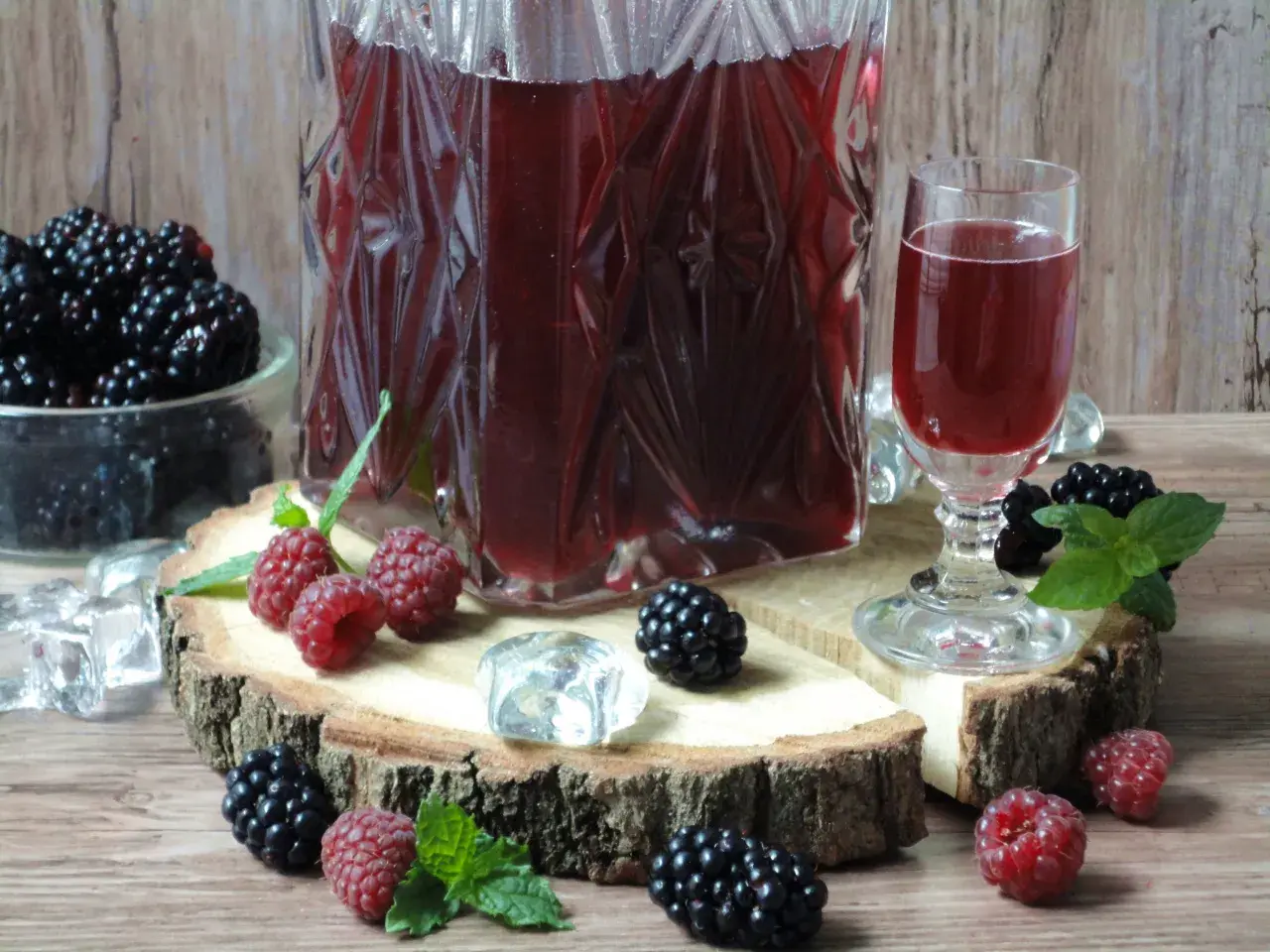 Aromatyczna nalewka ratafia w karafce i kieliszku, otoczona świeżymi malinami, jeżynami i kostkami lodu.