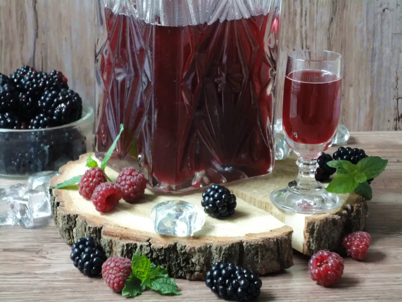Aromatyczna nalewka ratafia w karafce i kieliszku, otoczona świeżymi malinami, jeżynami i kostkami lodu.