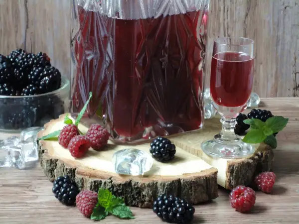 Aromatyczna nalewka ratafia w karafce i kieliszku, otoczona świeżymi malinami, jeżynami i kostkami lodu.
